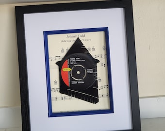 May include: A framed piece of sheet music with a black vinyl record cut out in the shape of a house. The record is from the song "Theme from Z-Cars" by Johnny Todd. The sheet music is for the song "Theme from Z-Cars" by Johnny Keating.