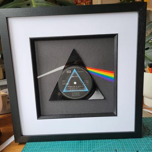 Puede incluir: Un álbum de disco enmarcado con un diseño de triángulo negro y una franja de arcoíris. El álbum se titula "The Dark Side of the Moon" de Pink Floyd.