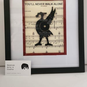 May include: A framed piece of art featuring a black vinyl record cut into the shape of a person, with the words "You'll Never Walk Alone" from the musical Carousel. The record is placed on top of sheet music with the same title. The frame is black with a red mount.