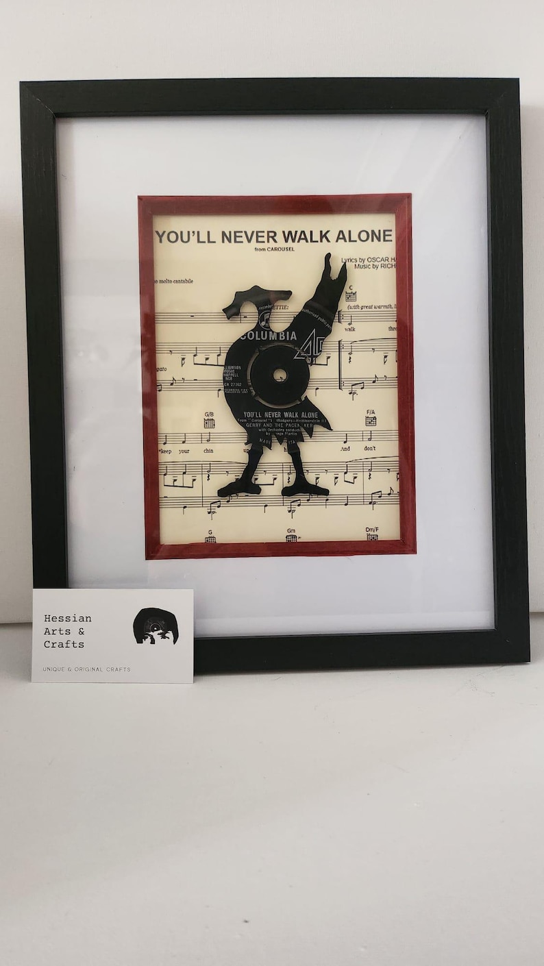 May include: A framed piece of art featuring a black vinyl record cut into the shape of a person, with the words "You'll Never Walk Alone" from the musical Carousel. The record is placed on top of sheet music with the same title. The frame is black with a red mount.