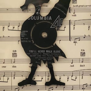 May include: A black vinyl record cut out in the shape of a person walking, superimposed on a sheet of music with the song title "You'll Never Walk Alone" printed on it. The record label is visible and says "Columbia" and "CA 27152".