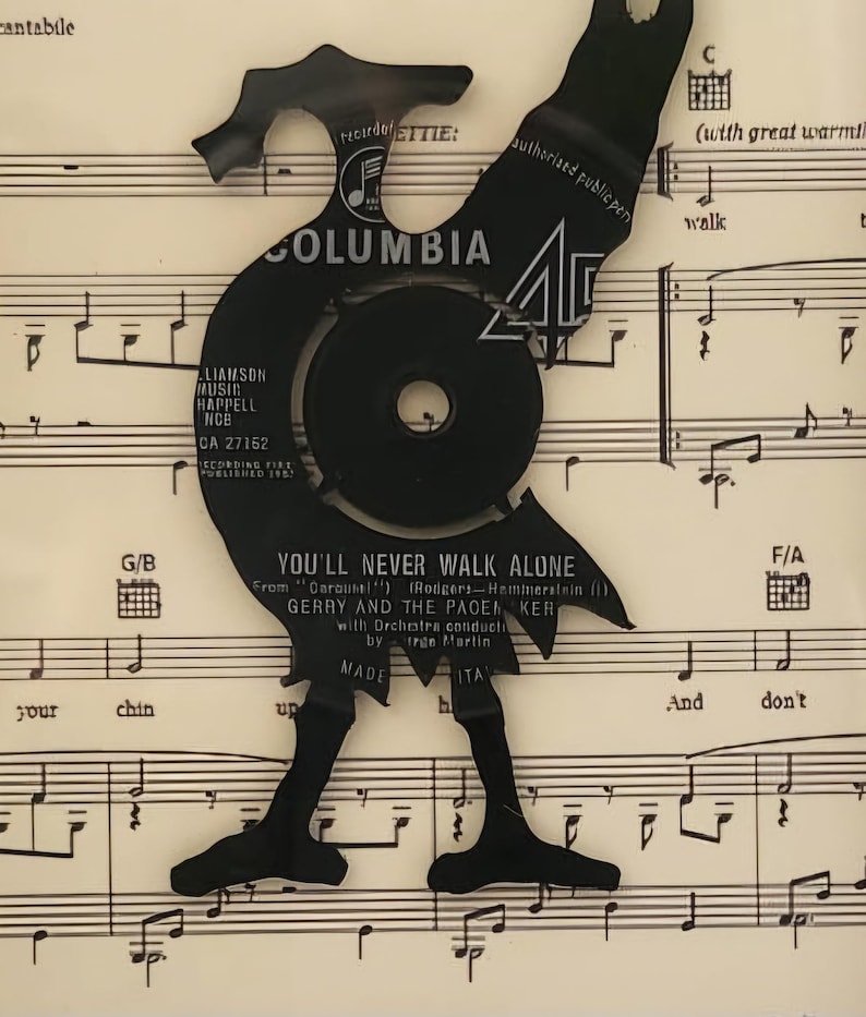May include: A black vinyl record cut out in the shape of a person walking, superimposed on a sheet of music with the song title "You'll Never Walk Alone" printed on it. The record label is visible and says "Columbia" and "CA 27152".