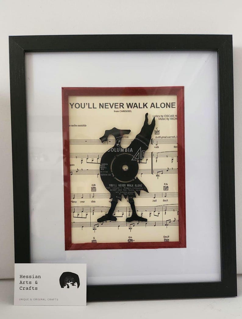May include: A framed black and white image of a vinyl record cut into the shape of a bird, with the words "You'll Never Walk Alone" from the musical "Carousel" printed on the background. The record is set against a red background and a sheet of music.