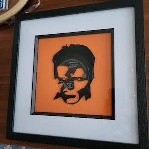 Könnte beinhalten: Eine schwarz-weiße Silhouette eines menschlichen Gesichts, aus einer Schallplatte ausgeschnitten, gerahmt in einem schwarzen Rahmen mit einem weißen Passepartout und einem orangenen Hintergrund. Das Plattenlabel ist im Auge der Silhouette sichtbar und zeigt den Text "Bob Dylan - Highway 61 Revisited".