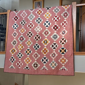 Puede incluir: Un edred&oacute;n hecho a mano con un patr&oacute;n de diamantes repetido. Cada diamante presenta un dise&ntilde;o de cruz en varios colores, incluyendo rojo, azul, amarillo y marr&oacute;n, sobre un fondo rosa con un peque&ntilde;o patr&oacute;n de puntos blancos. El edred&oacute;n se exhibe en interiores.
