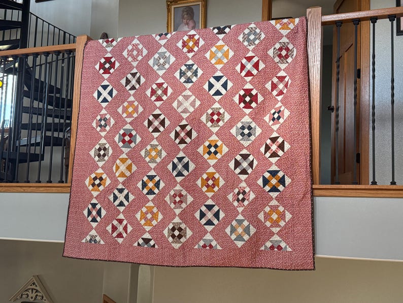 Puede incluir: Un edred&oacute;n hecho a mano con un patr&oacute;n de diamantes repetido. Cada diamante presenta un dise&ntilde;o de cruz en varios colores, incluyendo rojo, azul, amarillo y marr&oacute;n, sobre un fondo rosa con un peque&ntilde;o patr&oacute;n de puntos blancos. El edred&oacute;n se exhibe en interiores.