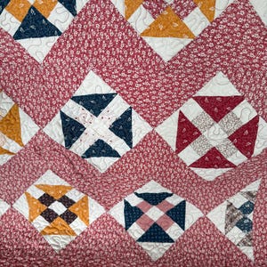 Puede incluir: Un textil acolchado con un patr&oacute;n repetitivo de formas geom&eacute;tricas en tonos de rojo, blanco, azul marino y dorado. El edred&oacute;n tiene un fondo con estampado floral y bloques en forma de diamante con costuras intrincadas, creando un dise&ntilde;o acogedor y decorativo.