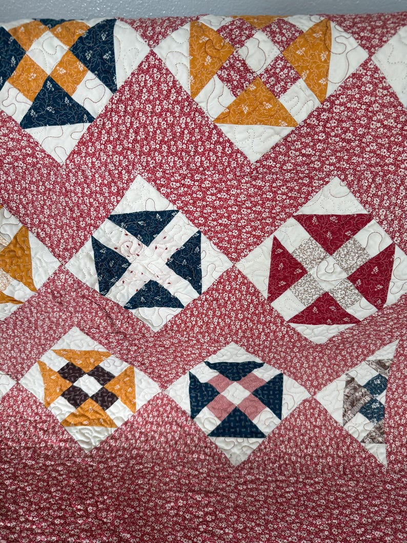 Puede incluir: Un textil acolchado con un patr&oacute;n repetitivo de formas geom&eacute;tricas en tonos de rojo, blanco, azul marino y dorado. El edred&oacute;n tiene un fondo con estampado floral y bloques en forma de diamante con costuras intrincadas, creando un dise&ntilde;o acogedor y decorativo.