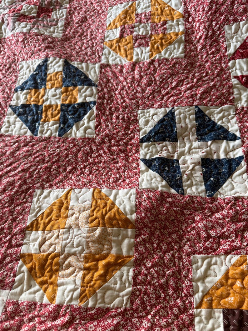 Puede incluir: Primer plano de un edred&oacute;n hecho a mano con un fondo floral rojo. El edred&oacute;n presenta bloques cuadrados con patrones de diamantes en tonos azul marino, dorado y blanco. El edred&oacute;n est&aacute; hecho de tela suave y tiene una textura acolchada.