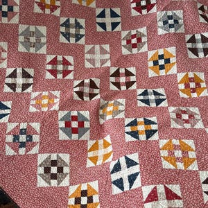 Puede incluir: Un edred&oacute;n hecho a mano con un patr&oacute;n repetitivo de bloques cuadrados. Cada bloque presenta un dise&ntilde;o central en varios colores, incluyendo rojo, azul, marr&oacute;n y amarillo, sobre un fondo blanco. La base del edred&oacute;n es una tela floral rosa.
