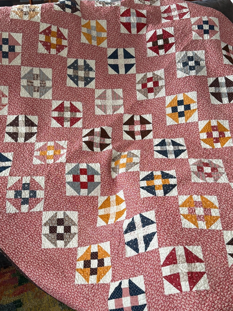 Puede incluir: Un edred&oacute;n hecho a mano con un patr&oacute;n repetitivo de bloques cuadrados. Cada bloque presenta un dise&ntilde;o central en varios colores, incluyendo rojo, azul, marr&oacute;n y amarillo, sobre un fondo blanco. La base del edred&oacute;n es una tela floral rosa.
