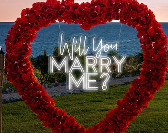 May include: A romantic outdoor proposal setup featuring a large heart-shaped floral arrangement in red. The heart frames the words "Will You Marry Me?" in neon. A red carpet leads to the heart, with the text "Kingdom Neon Art" and a heart symbol.