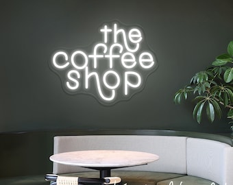May include: White neon sign with the text "the coffee shop" in a curved font. The sign is mounted on a dark green wall above a booth seating area with a round table and chair. The text "Kingdom Neon Art" is written in cursive on the booth.