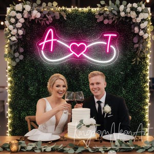 May include: A wedding celebration with a couple raising wine glasses in a toast. A two-tiered white cake, decorated with flowers, is on the table. A neon sign with the letters "A" and "T" and a heart shape is visible. The backdrop is a green hedge wall.