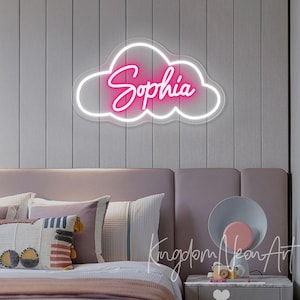 May include: A pink neon sign in the shape of a cloud with the name "Sophia" written in a cursive font. The sign is mounted on a white wall above a bed with pillows and a bedside table.