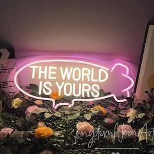 Puede incluir: Un letrero de neón en un contorno rosa, con forma de bocadillo, muestra el texto "THE WORLD IS YOURS" en blanco. El letrero está rodeado de vegetación, flores rosas y amarillas y una claqueta de cine.