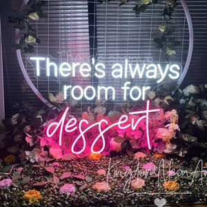 May include: A white neon sign with the text "There's always room for dessert" in pink neon letters. The sign is surrounded by pink and white flowers.