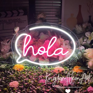 Op de afbeelding: Een neonbord in de vorm van een tekstballon met het woord "hola" in roze. Het bord heeft een witte omtrek en staat tegen een bloemenachtergrond. Het bord is van Kingdom Neon Art.