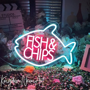 Op de afbeelding: Een neonbord in de vorm van een vis met de woorden "Fish & Chips" in roze neonletters. Het bord staat op een bed van groene en roze bloemen.