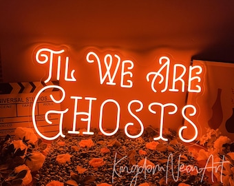 May include: A bright orange neon sign displaying the text "TIL WE ARE GHOSTS" in a stylized cursive font. The sign is mounted on a clear acrylic backing and is positioned above a bed of artificial orange flowers and dark green foliage.