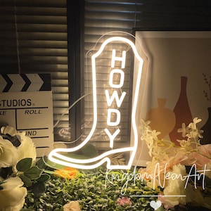 以下が含まれることがあります： カウボーイブーツの形をした白いネオンサインで、ブーツの中に「HOWDY」という文字が縦に書かれています。サインは点灯しており、緑と白の植物のベッドの上に置かれています。