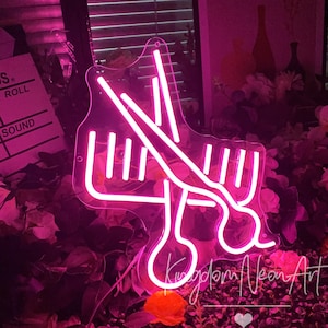 Puede incluir: Un letrero de neón rosa que representa un par de tijeras y un peine, un símbolo común de las peluquerías. El letrero está iluminado y tiene un contorno blanco. El letrero está sobre un fondo de flores rosas.