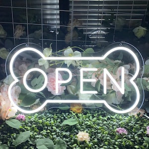 以下が含まれることがあります： 骨の形をした白いネオンサインで、中央に「OPEN」の文字があります。サインは、ピンクの花が咲いた緑の茂みの前にあります。