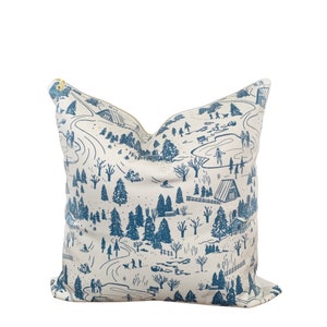 May include: A decorative pillow with a winter scene design. The pillow features a cream-colored background with a blue illustration of a snowy landscape, including trees, houses, and people sledding. The pillow has a gold zipper.