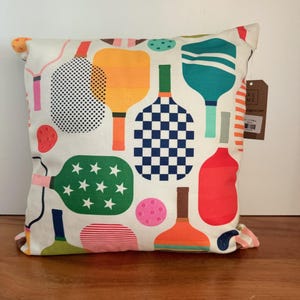 May include: Square decorative pillow with a white background and a colorful pattern of pickleball paddles and balls. The paddles are in various colors, including orange, green, blue, and red, with different designs.