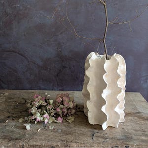 Peut inclure: Un grand vase en céramique blanc cassé avec une texture verticale ondulée et plissée. Une fine branche brune et nue dépasse de l'ouverture supérieure du vase. À côté du vase se trouve un petit bouquet de fleurs d'hortensia séchées rose pâle.