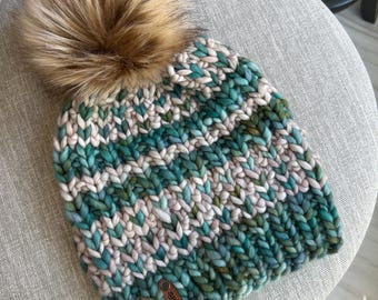 May include: A chunky-knit beanie hat in teal, cream, and olive green, with a brown faux fur pom-pom. The hat has a textured design and a small leather tag that reads "SJK Knits".