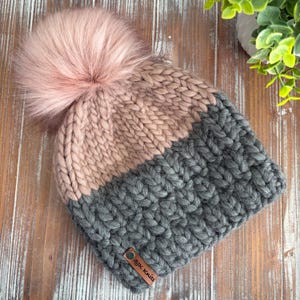 May include: A chunky knit beanie in gray and blush pink with a large, fluffy pink pom-pom. The hat features a leather tag with the text "SJK Knits". This warm winter hat is perfect for cold weather.