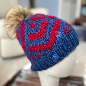 May include: A blue and red knitted beanie with a faux fur pom pom on top.
