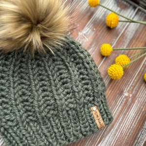 May include: A dark green knitted beanie with a fluffy brown pom pom on top. The beanie has a brown leather tag with the text "SIK KNITS" on it.