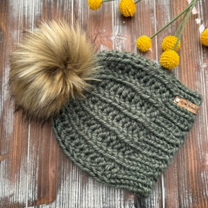 May include: A green knitted beanie with a brown faux fur pom pom. The beanie has a brown leather tag with the text "SIK Knits" on it.