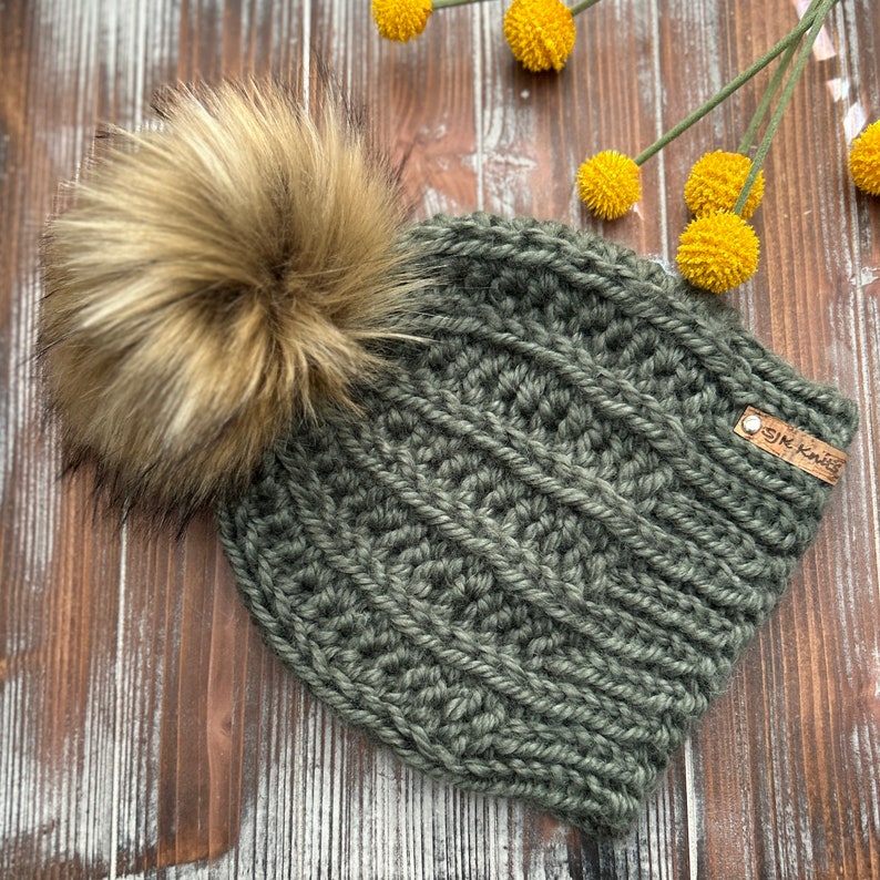 May include: A green knitted beanie with a brown faux fur pom pom. The beanie has a brown leather tag with the text "SIK Knits" on it.