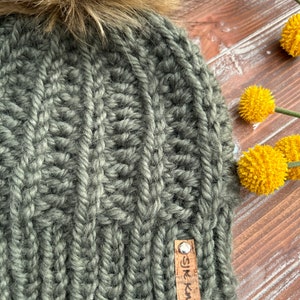 May include: A dark green knitted beanie with a faux fur pom pom on top. The beanie has a cable knit pattern and a brown leather tag that says "SJK Knits".