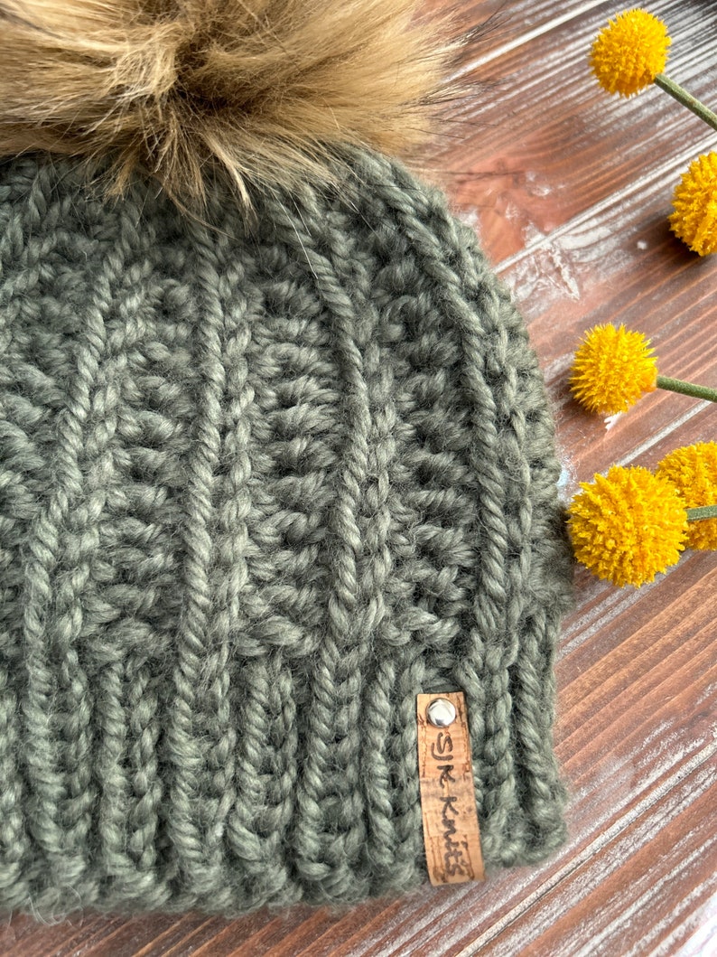 May include: A dark green knitted beanie with a faux fur pom pom on top. The beanie has a cable knit pattern and a brown leather tag that says "SJK Knits".