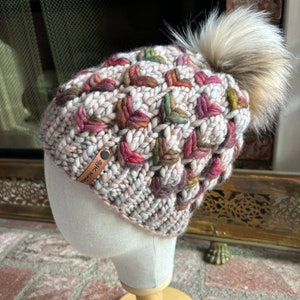 May include: A beige knitted hat with a colorful geometric pattern and a faux fur pom pom. The hat has a leather tag with the text "Silk Road" on it.
