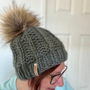 May include: A dark green knitted beanie with a brown faux fur pom pom. The beanie has a woven label that says "Stitch & Soul".