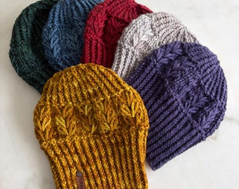 May include: A collection of six chunky knit beanies in mustard yellow, dark purple, light grey, deep red, blue, and dark green. Each hat features a thick ribbed brim and a textured cable knit pattern. A small brown leather tag labelled SJKnits is attached.