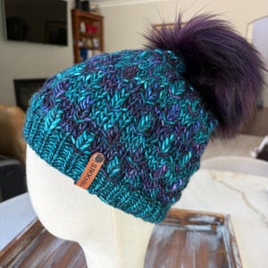 May include: A hand-knit beanie featuring a variegated yarn pattern in shades of teal, blue, and purple. The hat has a ribbed brim, a textured stitch design, and a large, fluffy dark purple pom-pom. A brown leather tag is attached with the text SJKknits.