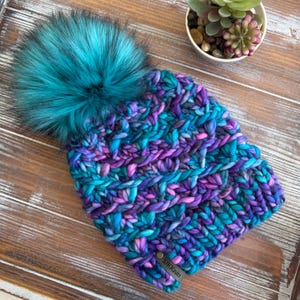 May include: A chunky knit beanie featuring a variegated yarn pattern in shades of teal, purple, and blue. The hat is topped with a large, fluffy teal faux fur pom-pom and includes a small rectangular brown tag with the text SJKKnits.