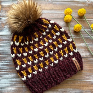 May include: A burgundy knit beanie with a white and mustard yellow chevron pattern. The beanie has a faux fur pom pom on top and a brown leather tag that says "sik knits".