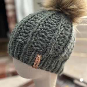 May include: A green knitted beanie with a faux fur pom pom and a brown leather tag that says "SK Knit".