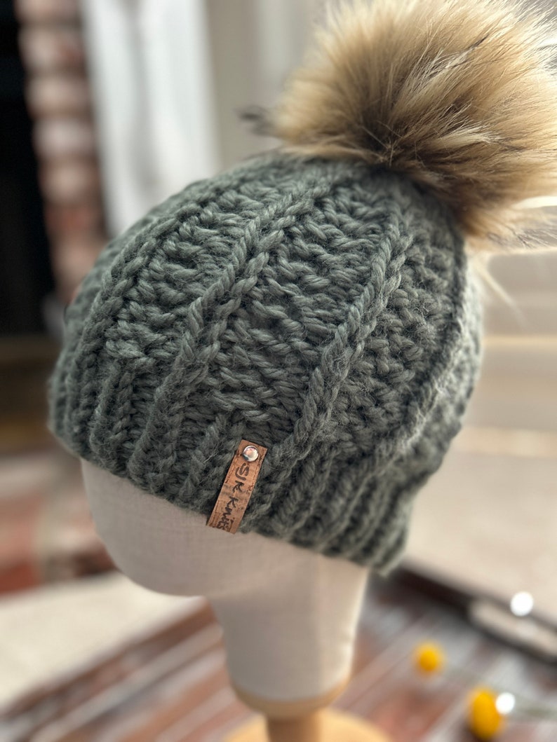 May include: A green knitted beanie with a faux fur pom pom and a brown leather tag that says "SK Knit".
