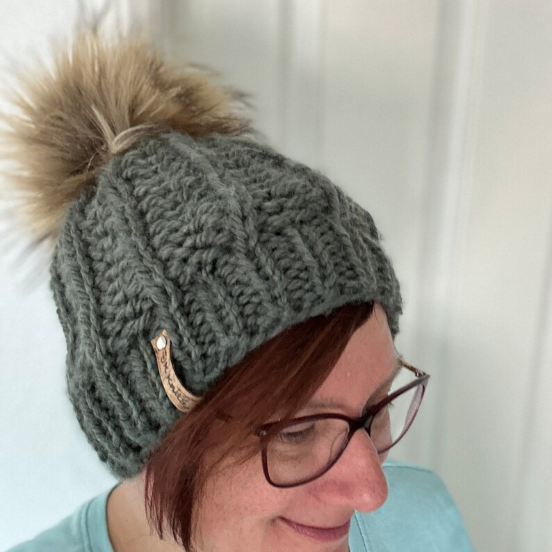 May include: A grey knitted beanie with a brown and white faux fur pom pom. The beanie has a brown leather tag with the word "Handmade" on it.