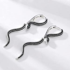 Puede incluir: Un par de pendientes de aro de plata con forma de serpiente, con un cuerpo curvado incrustado con pequeñas piedras negras. La superficie metálica lleva el sello S925, que indica plata de ley, y las cabezas de las serpientes tienen ojos verdes.