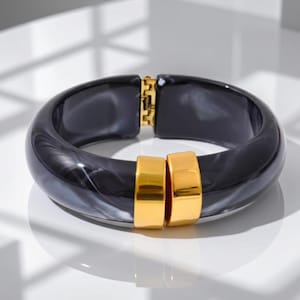 Może przedstawiać: Otwierana bransoleta typu bangle wykonana z polerowanej żywicy o czarno-szarym marmurkowym wzorze. Projekt zawiera dwa gładkie, prostokątne akcenty z metalu w kolorze złotym z przodu oraz zawias z metalu w kolorze złotym z tyłu.