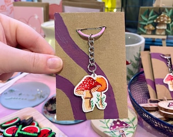 Puede incluir: Un llavero con un dije plano ilustrado de dos setas, una con sombrero rojo y puntos blancos y la otra con sombrero naranja, unido a una cadena de metal plateado y una anilla rosa, montado sobre una tarjeta de presentación de cartón marrón.